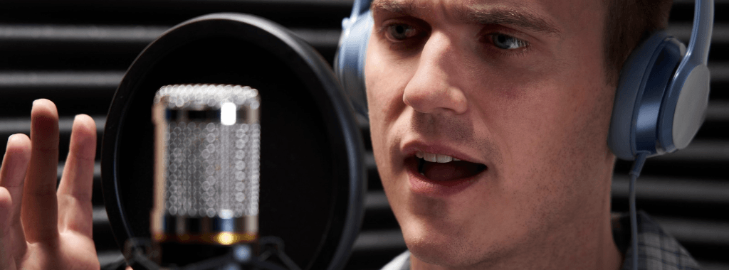 Close up of man wearing headphones in a vocal booth in front of a microphone with a pop filter and acoustic foam in the background