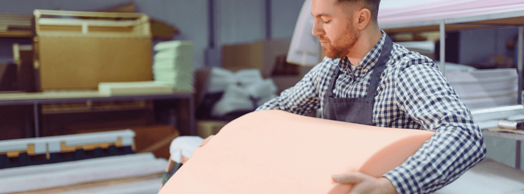 Caucasian man in a factory wearing a checked shirt and apron examining a light pink piece of seating foam