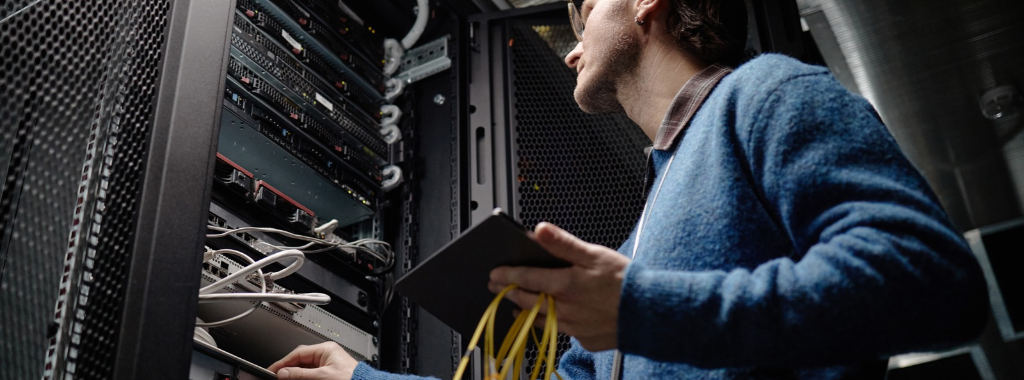 Man in a server room holding a yellow cable and a tablet. He is wearing a blue sweater and glasses and is working on a server rack.